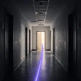 A dark office corridor with a single violet streak of light running down the floor to one lit doorway