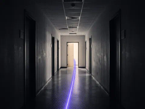 A dark office corridor with a single violet streak of light running down the floor to one lit doorway