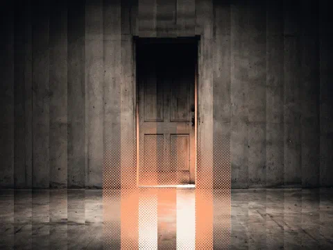 A tall wooden door in a concrete wall with a band of warm orange halftone light spilling from its lower gap