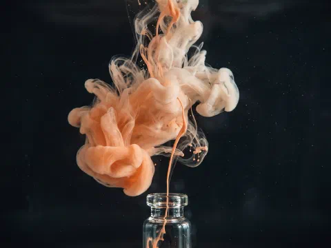 A thin thread of peach ink escaping a small glass vial and blooming into a cloud in dark water.