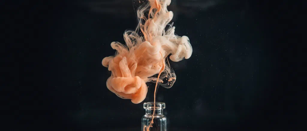 A thin thread of peach ink escaping a small glass vial and blooming into a cloud in dark water.
